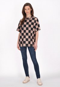 Blusa de gran tamaño con mangas cortas en un patrón a cuadros en negro y beige, fabricada en tejido suave. Combinada con jeans oscuros y zapatillas beige.