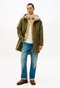 Wodoodporna oliwkowozielona parka z kapturem, beżowa koszula, kremowy sweter w warkocze, jasnoniebieskie dżinsy i brązowe skórzane buty.