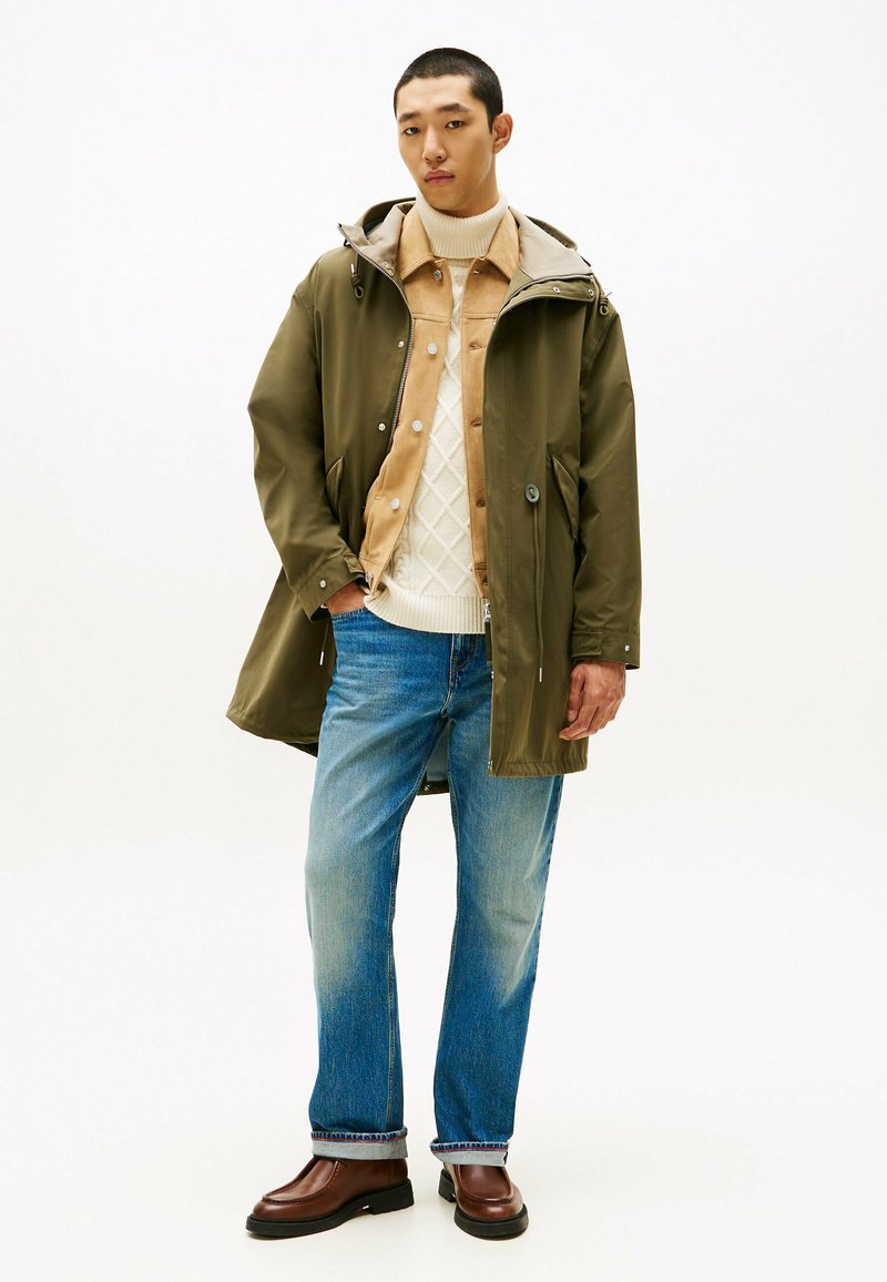 Wodoodporna oliwkowozielona parka z kapturem, beżowa koszula, kremowy sweter w warkocze, jasnoniebieskie dżinsy i brązowe skórzane buty.