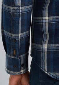 Blauw geruite blouse met lange mouwen en witte en lichterblauwe strepen, voorzien van knoopmanchetten en een klein label nabij de naad.