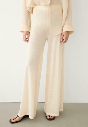 Person trägt weite, cremefarbene Hosen und eine transparente, langärmelige Bluse, dazu schwarze Flip-Flop-Sandalen.