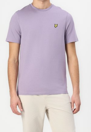 Mann trägt ein hellviolettes T-Shirt mit kurzen Ärmeln und Rundhalsausschnitt mit kleinem gelbem Vogel-Logo auf der linken Brust, kombiniert mit beigen Hosen.