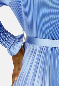 Abito plissettato azzurro chiaro con texture satinata, caratterizzato da maniche lunghe, polsini arricciati e un dettaglio in vita abbinato.