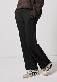 Pantaloni neri con un design a gamba dritta, caratterizzati da una texture liscia e strisce laterali a contrasto in una tonalità più scura. Indossati con sneaker chiare.