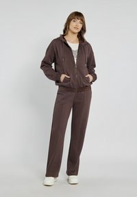 Brązowy hoodie z zamkiem błyskawicznym, sznurkami i bocznymi kieszeniami; w komplecie z pasującymi brązowymi spodniami. Miękka tkanina, luźny krój i elastyczny pas.