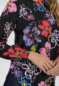 Blusa floreale in tessuto nero con fiori di un brillante rosa, rosso, blu e verde. Maniche lunghe con polsini arricciati; colletto visibile.