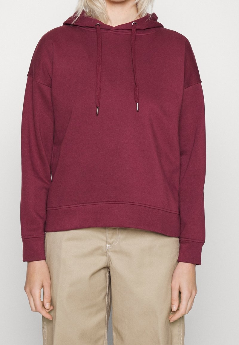 Personne portant un sweat à capuche bordeaux uni avec des cordons et un pantalon beige, visible des épaules aux hanches sur un fond uni.