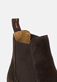 Bottine en daim marron avec des panneaux latéraux en mesh, des détails cousus et un intérieur en cuir marron. Dispose d'une languette de traction au talon.