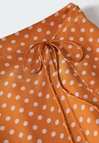 Blusa arancione a pois con finitura in satin, dettaglio di cravatta regolabile e collo rotondo. Il tessuto è leggero e liscio con pois bianchi.