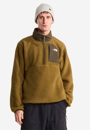 Mann trägt einen olivgrünen Fleecepullover mit Brusttasche und weißer Mütze, steht vor einem schlichten weißen Hintergrund.