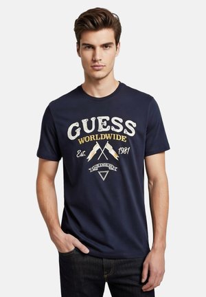 Hombre con camiseta azul marino de Guess con banderas cruzadas, texto "MUNDIAL", "Est. 1981" y "LOS ÁNGELES", con la mano en el bolsillo sobre fondo blanco.