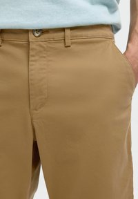 Bež pamukove chinos hlače ravnog kroja, s kopčanjem na gumb i bočnim džepovima. Glatka tekstura i uredna šavovi.