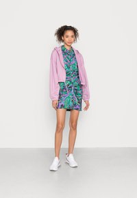 Hoodie curto roxo por cima de um vestido com padrão verde e azul com franzido, combinado com tênis brancos, em um fundo simples.