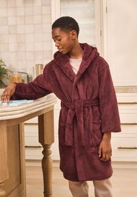 Accappatoio in pile color bordeaux con cappuccio, cintura in vita e tasche laterali. Il tessuto presenta una superficie morbida e strutturata. Posizionato su un bancone da cucina.
