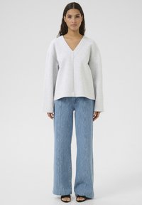 Lys grå V-hals sweatshirt med lange ærmer, parret med blå, bredbenede, teksturerede bukser. Sorte, åbne sandaler fuldender outfittet.