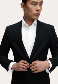 Mann, der einen Knopf an einem schwarzen Anzugjackett über einem weißen Hemd schließt, während er seitlich gegen einen schlichten Hintergrund blickt.