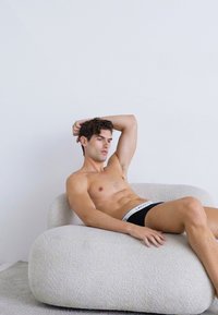 Krągłe, off-white tapicerowane krzesło z teksturowanym materiałem. Bez koszulki mężczyzna siedzi swobodnie, ubrany w czarne majtki z białą gumką.