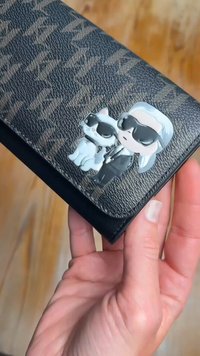 Juodas tekstūruotas piniginė su blizgiu emalio smeigtuku, vaizduojančiu personažą su sidabriniais plaukais ir katę, abu dėvinčius akinius nuo saulės.