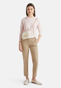 Helles rosa gestricktes Sweatshirt, beige maßgeschneiderte Hose, cremefarbige Handtasche mit goldenem Zubehör und beigen Loafers in einer stehenden Pose vor einem weißen Hintergrund.