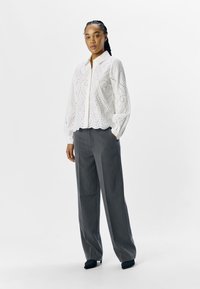 Blouse blanche cropped avec un motif à œillets et un collier ; associée à un pantalon gris taille haute ; le modèle se tient dans des chaussures noires plates.
