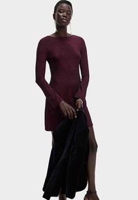 Maroon long-sleeve dress with a fitted silhouette, round neckline, and bell-shaped cuffs. Model holds a black jacket, showcasing smooth texture.