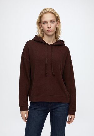 Jonge vrouw met blond haar die een donkerbruine hoodie en blauwe spijkerbroek draagt, staand tegen een effen lichte achtergrond.