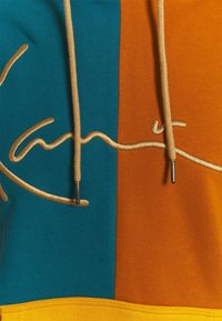 Een hoodie met turquoise en oranje panelen, voorzien van gouden borduursels en beige trekkoorden. Zachte stof met een gestructureerde afwerking.