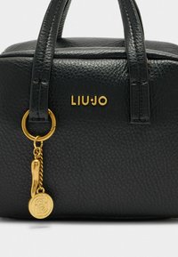 Sac à main en cuir texturé noir avec des métaux dorés, présentant un logo de marque et un charme décoratif sur une chaîne. Forme compacte et rectangulaire.