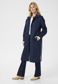 Marineblauwe trenchcoat tot de knie met een kraag, vijf knopen en twee zijzakken. Gecombineerd met flared jeans en beige enkellaarsjes.