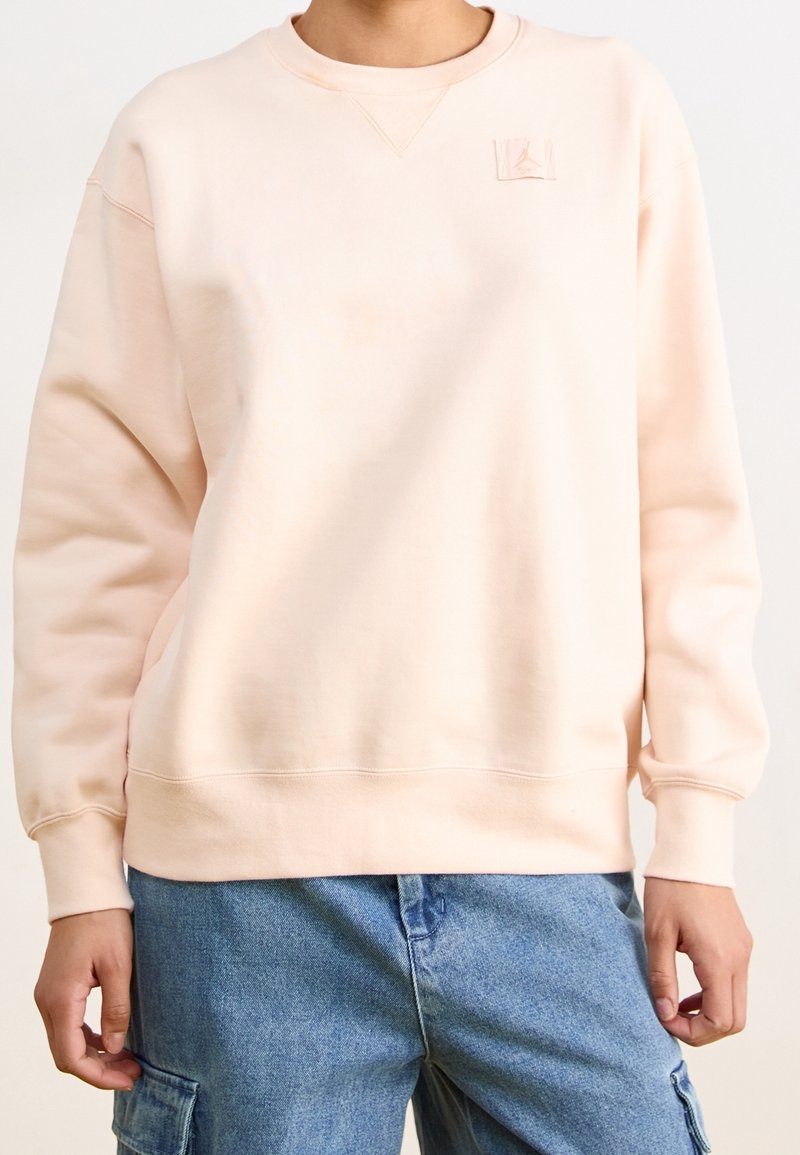 Personne portant un sweat-shirt à manches longues rose pâle avec un petit logo près de l'encolure et un pantalon cargo en denim bleu.