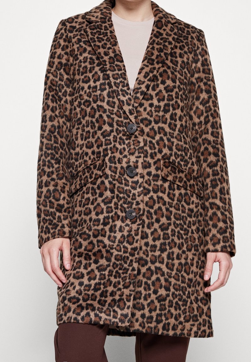 Personne portant un manteau mi-long à imprimé léopard marron et noir avec trois boutons, par-dessus un haut beige clair et un pantalon marron foncé.