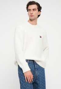 Weißer Strickpullover mit Rundhalsausschnitt und gerippten Bündchen, versehen mit einem kleinen Logopatch. Kombiniert mit blauen Jeans aus Denim, die mit Textmustern versehen sind.