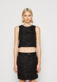 Svart crop top med rynkdetalj, i kombination med en svart kjol med fickor, båda i en slät, glänsande textil. Enkla, rena linjer.
