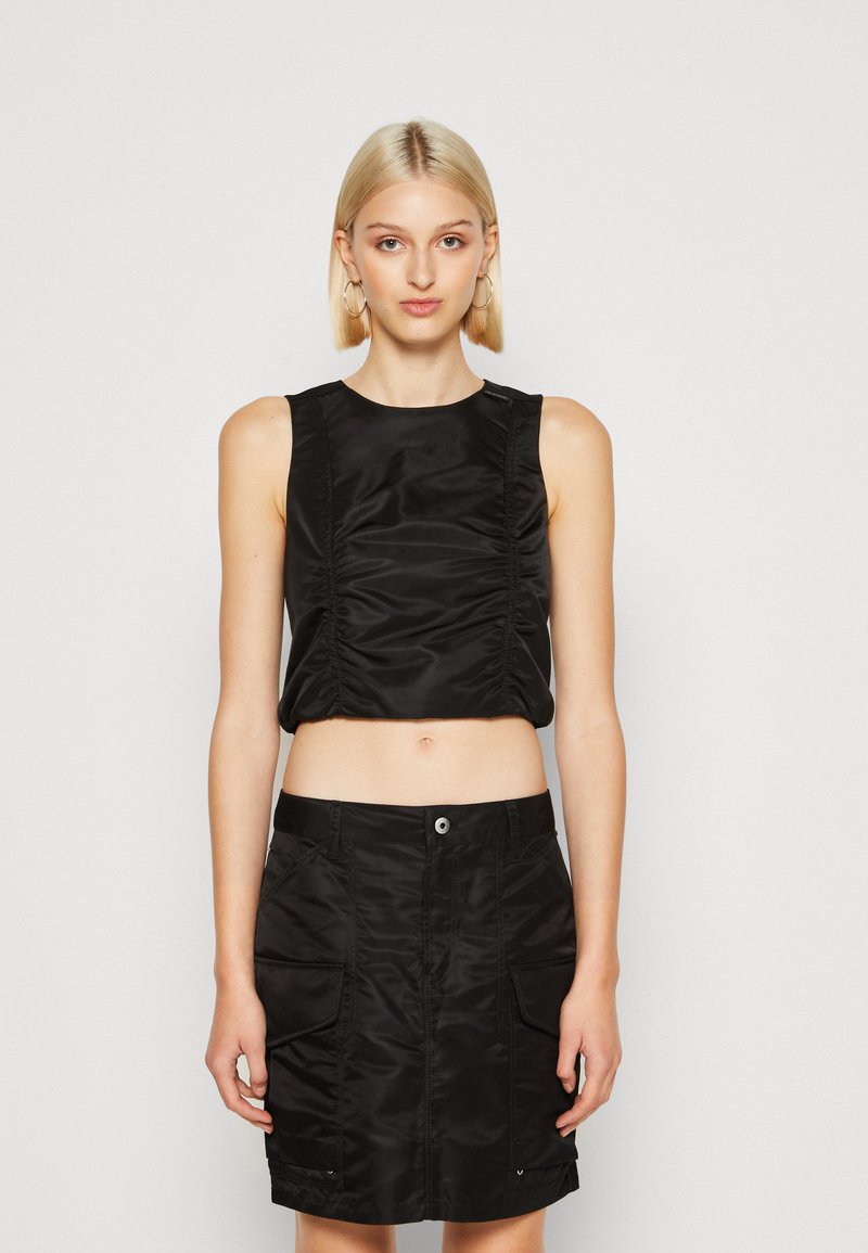 Svart crop top med rynkdetalj, i kombination med en svart kjol med fickor, båda i en slät, glänsande textil. Enkla, rena linjer.