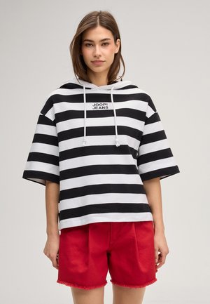 Jonge vrouw draagt een zwart-wit gestreepte hoodie met de tekst "JOOP! JEANS" en rode korte broek, staande tegen een effen lichte achtergrond.