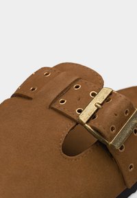 Πίσω λουράκι από καφέ σουέντ πέδιλο με αγκράφα από μέταλλο με χρυσό χρώμα και πολλαπλές τρύπες σε απλό φόντο.