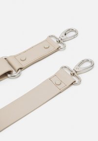 Beige neplederen band met zilveren hardware en klinknagels, voorzien van drukknophaken voor bevestiging en een gladde textuur.