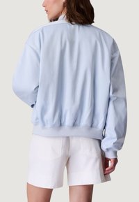 Jasnoniebieska kurtka bomberka z okrągłym dekoltem, elastycznymi mankietami i pasem. Gładka faktura, luźny krój, widziana od tyłu. Widoczne białe szorty.