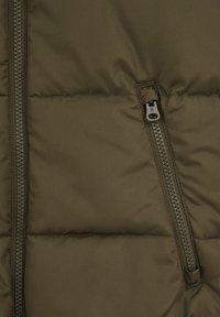 Olivgrön quiltad puffjacka med en metallblixtlås på vänster sida. Slät textur med vadderade sektioner för isolering.