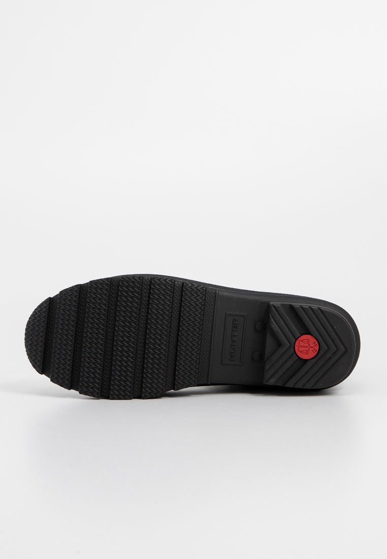 Suela de zapato de goma negra con patrones de relieve profundos y un pequeño logo circular rojo cerca del talón, sobre un fondo blanco.