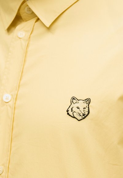 Κίτρινο πουκάμισο με κουμπιά και μικρό κεντητό μπαλώμα με κεφάλι αλεπούς στην αριστερή περιοχή του στήθους.
