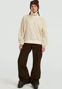 Donna che indossa un pullover in pile beige, pantaloni cargo marrone scuro, scarpe nere e un berretto marrone, in piedi davanti a uno sfondo semplice.