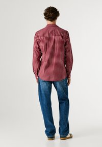 Camicia a maniche lunghe a quadri rossi e bianchi con colletto abbottonato, abbinata a jeans in denim blu. Cuciture visibili e dettagli di vestibilità su misura.