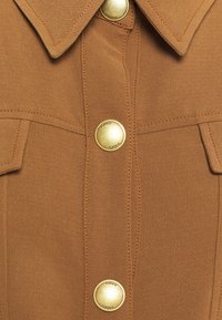 Camicia marrone con un design strutturato, ampio colletto e bottoni dorati. Tessuto testurizzato con cuciture visibili e dettagli delle tasche.