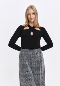 Czarna bluzka z długim rękawem, z wycięciami na ramionach i dekoltem w kształcie klucza, zestawiona z szarymi szerokimi spodniami z fałdami, obszytymi pionowymi srebrnymi paskami.