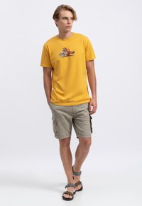 T-shirt en coton jaune avec un imprimé graphique sur la poitrine, associé à un short cargo beige de longueur genou et des sandales noires.
