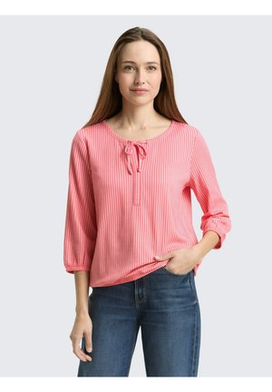 Kobieta w różowej bluzce w pionowe paski z wiązanym dekoltem i rękawami do łokcia, w połączeniu z niebieskimi jeansami, pozująca z jedną ręką w kieszeni.