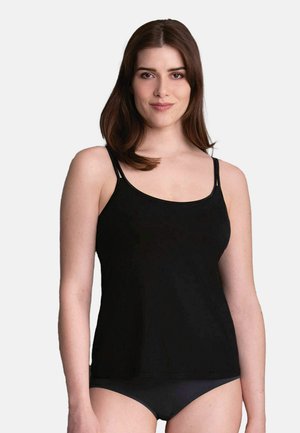 Junge Frau mit glatten braunen Haaren, die ein schwarzes Camisole-Top und schwarze Unterwäsche trägt, leicht lächelnd, vor einem schlichten hellen Hintergrund.