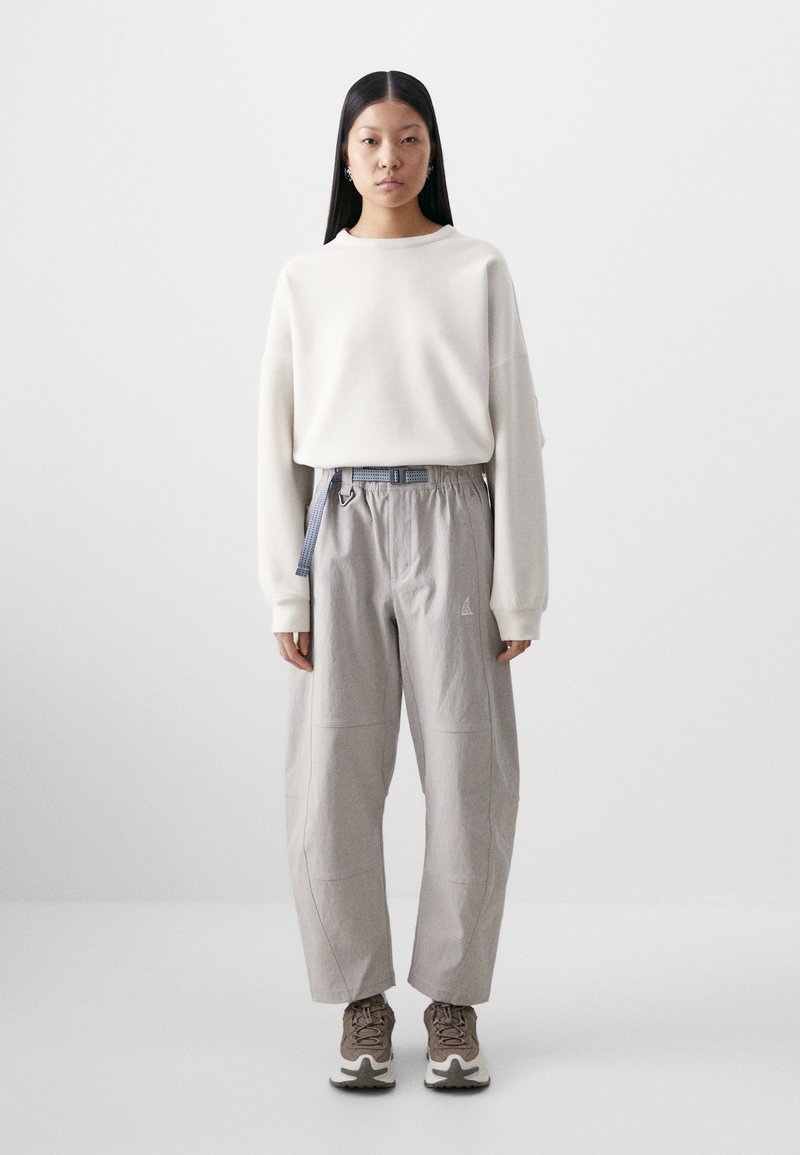 Light beige sweatshirt with drop shoulders, paired with loose gray cargo pants featuring an elastic waistband and a blue patterned belt, and sneakers.