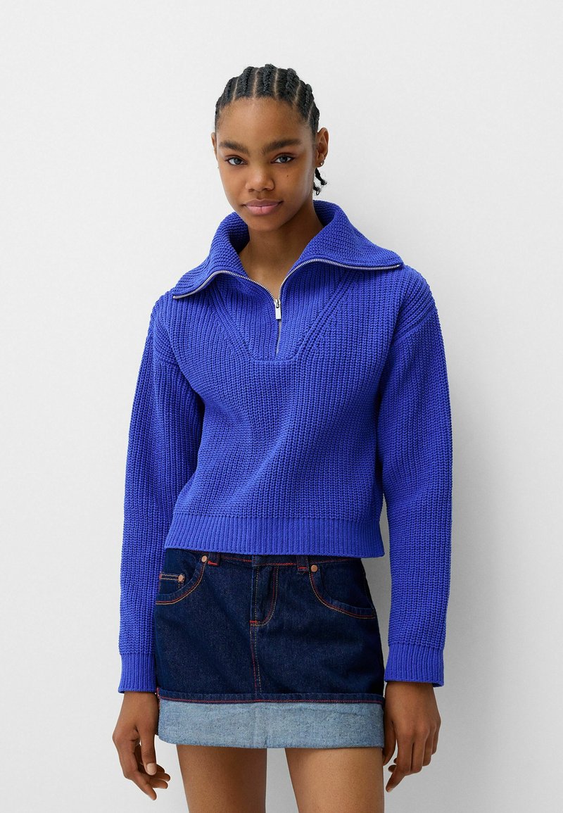 Pullover lavorato a maglia viola con colletto alto e chiusura a mezza zip; motivo testurizzato, vestibilità cropped, abbinato a una mini gonna di jeans.